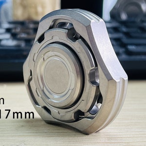 May include: A silver titanium fidget spinner with a textured outer ring and a smooth inner ring. The spinner is 48x48x17mm and weighs 70g.