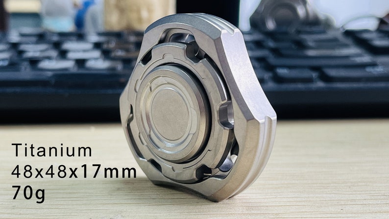 May include: A silver titanium fidget spinner with a textured outer ring and a smooth inner ring. The spinner is 48x48x17mm and weighs 70g.