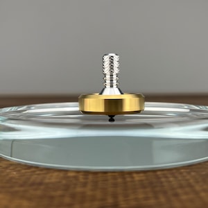 May include: A spinning top made of brass and silver metal spinning on a clear glass surface.