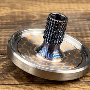 May include: A metal spinning top featuring a circular base with a copper-colored honeycomb pattern inlay. The central stem has a textured, knurled grip and a heat-treated, iridescent blue and purple finish, surrounded by a smooth, polished silver-toned rim.