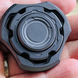 May include: A black metal fidget spinner with a circular design and multiple rings. The spinner has a textured surface and is held in a hand.