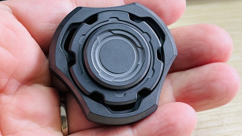 May include: A black metal fidget spinner with a circular design and multiple rings. The spinner has a textured surface and is held in a hand.