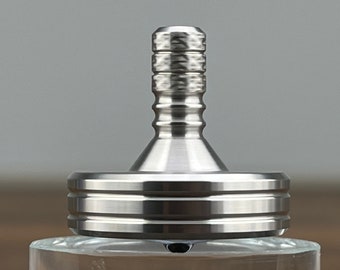 May include: A silver metal spinning top with a textured top and a wide base. The top is spinning on a clear glass surface.