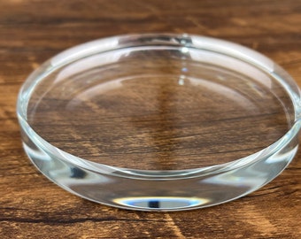 May include: A clear glass display stand with a round, slightly raised edge. The stand is perfect for showcasing small items like jewelry, figurines, or candles.