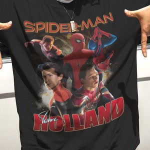 Może przedstawiać: Czarny T-shirt z grafiką Spider-Mana i Toma Hollanda. Tekst "Spider-Man" jest w żółtych i czerwonych literach, a tekst "Tom Holland" jest w czerwonych literach.