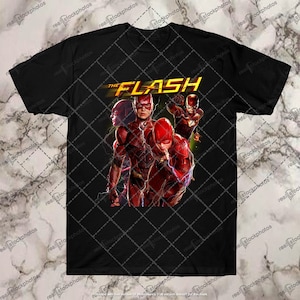 Może przedstawiać: Czarny T-shirt z nadrukiem graficznym Flasha, superbohatera z czerwonymi i żółtymi błyskawicami. Tekst "THE FLASH" jest nadrukowany na żółto nad obrazkiem.
