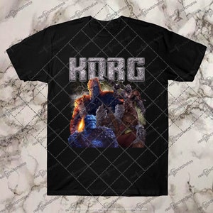 Peut inclure: T-shirt noir avec le mot "KORG" en argenté texturé. En dessous, un graphisme de plusieurs personnages bleus, marron et orange. Le t-shirt est fait d'une matière douce.