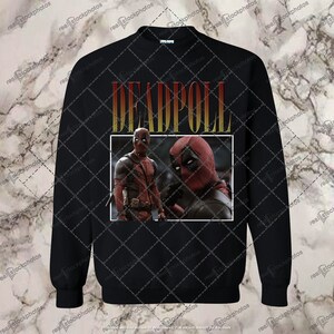 K&ouml;nnte beinhalten: Schwarzes Sweatshirt mit einem Grafikdruck von Deadpool in einem gelben und roten 'DEADPOOL'-Textfeld.