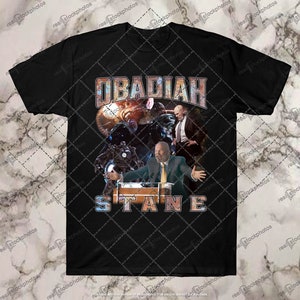 May include: Black t-shirt with a graphic print of a man in a suit standing in front of a large, metallic robot. The text "OBADIAH STANE" is printed in a distressed, vintage style.