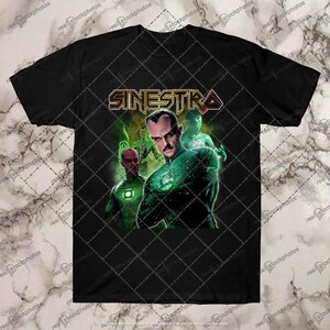 Op de afbeelding: Zwart T-shirt met een afbeelding van een superheldenfiguur met groene en gele accenten. De tekst "SINESTRA" staat in gouden letters boven het figuur.