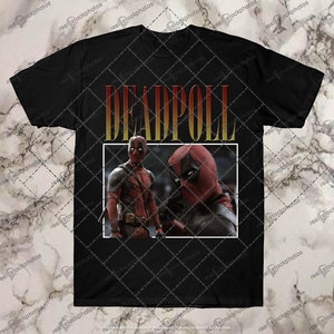 K&ouml;nnte beinhalten: Schwarzes T-Shirt mit einem Aufdruck von zwei Deadpool-Charakteren vor einem roten und gelben Hintergrund. Der Text "DEADPOOL" ist in gelben Buchstaben mit rotem Umriss gedruckt.