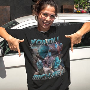 May include: Black t-shirt with a vintage-style graphic of Yondu from Guardians of the Galaxy. The graphic features Yondu and other characters from the movie, with the text "Yondu" and "Guardians of the Galaxy" in a bold, colorful font. The shirt also includes the signature "Rooster Michael".