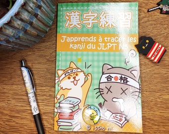May include: A green and white book with the title "漢字練習 J'apprends à tracer les kanji du JLPT N5" and illustrations of two cartoon animals. The book is open to a page with a cartoon dog wearing a headband that says "がんばって!" and a cartoon cat wearing a headband that says "合格" with two X's under it. The book is on a wooden surface.