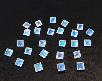 Könnte beinhalten: Eine Ansammlung quadratisch geschliffener, schillernder Mondstein-Edelsteine. Jeder Stein zeigt ein Farbspiel aus Blau und Weiß, verstreut auf dunklem Untergrund. Geeignet für Schmuckherstellung oder Sammler.