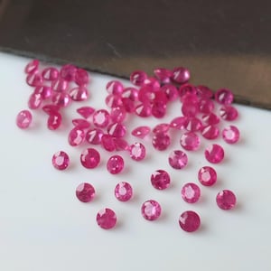 May include: A collection of small, round, faceted ruby gemstones. The rubies are a vibrant pinkish-red color and are scattered across a white surface. The gemstones vary slightly in size and shape, with a few oval-cut stones.