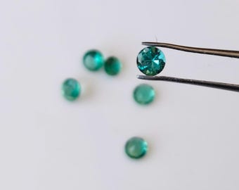 May include: Five round, faceted, green gemstones. One gemstone is held by tweezers, the others are scattered on a white surface.