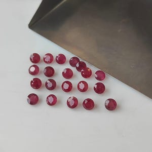 May include: A collection of faceted, round, deep red gemstones. The gems are scattered on a white surface near a silver metal tray. The gemstones have a polished, reflective surface, and are likely rubies.