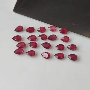 May include: A collection of twenty-one pear-shaped, deep red gemstones. The gems are arranged on a white surface, with a dark gray metal tray in the background. The gemstones are faceted and appear to be polished.