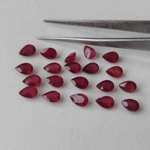 May include: A selection of deep red, pear-shaped gemstones. The faceted stones are arranged on a white surface, with silver tweezers in the background. The gems have a polished, reflective surface.