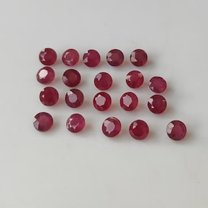 Puede incluir: Una colección de gemas redondas, facetadas, de color rojo intenso, posiblemente rubíes, dispuestas sobre una superficie blanca. Las gemas tienen una superficie pulida y reflectante, y varían ligeramente en color y tamaño. La disposición es dispersa, sin patrón aparente.