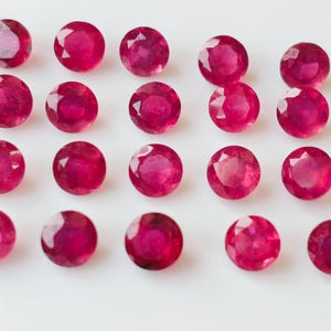 May include: Twenty round, red gemstones arranged in rows on a white surface.