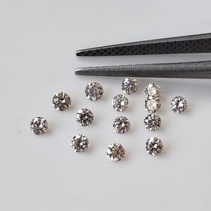 May include: A collection of thirteen small, round, brilliant-cut loose diamonds with a clear, sparkling appearance. The stones are arranged in a loose cluster, with a pair of dark grey metal tweezers positioned at the top of the frame.