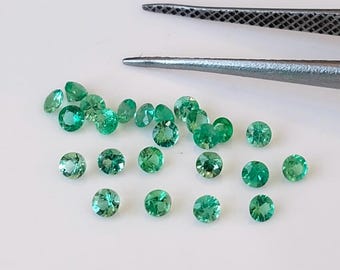 May include: A collection of round, green gemstones, likely emeralds, scattered on a white surface. The gemstones are various sizes and have a slight sparkle.
