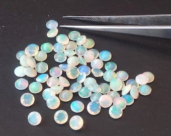 Pode incluir: Uma coleção de pequenas opalas redondas e facetadas em tons de branco, azul e verde. As pedras preciosas estão espalhadas sobre uma superfície escura, com um par de pinças prateadas no fundo.