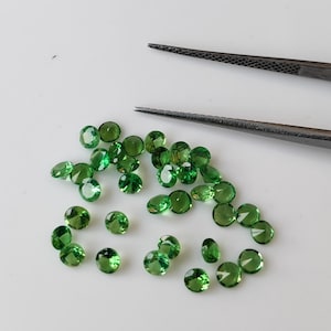 May include: A collection of round, green gemstones, likely peridot, scattered on a white surface. The gemstones are various sizes and have a smooth, polished finish.