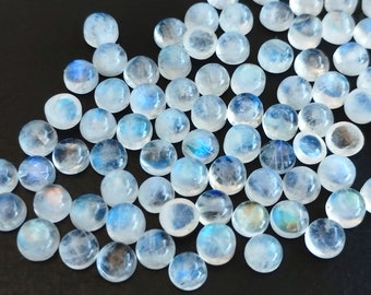 Puede incluir: Un conjunto de cabujones redondos de piedra lunar pulidos. Las piedras son translúcidas con una base blanca lechosa y exhiben una adularescencia azul. Los cabujones están dispersos sobre una superficie oscura, mostrando su cualidad iridiscente.
