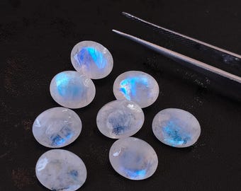 May include: Eight oval-shaped moonstone cabochons with iridescent blue and white colors. The stones are arranged in a semi-circle on a black surface.