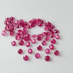 Può includere: Una collezione di pietre preziose di rubino rosa sciolte, sparse su una superficie bianca. Le pietre rotonde e sfaccettate sono disposte a forma di cuore.