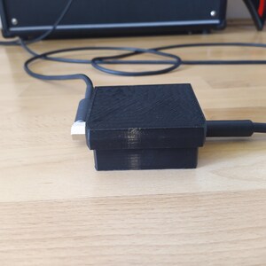Könnte beinhalten: Eine schwarze, im 3D-Druck hergestellte Box mit einem schwarzen Kabel, das eingesteckt ist. Die Box steht auf einer Holzoberfläche.