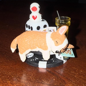 Pode incluir: Uma figura peculiar com um corgi adormecido com pelo castanho e branco, descansando sobre uma ficha de poker preta e branca. Um dado, um copo de shot, cartas de baralho e uma nota de cem dólares completam a cena.