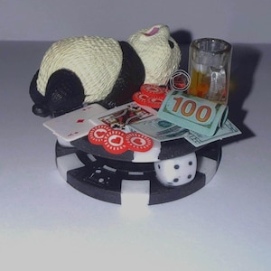 Pode incluir: Uma figura de panda preto e branco em cima de uma ficha de poker preta e branca, rodeada por cartas de jogar, fichas vermelhas, um copo de shot, um dado e uma nota de 100 dólares enrolada.