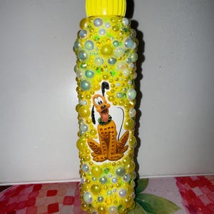 May include: A yellow water bottle decorated with yellow and white beads. The bottle features a cartoon image of Pluto. The bottle has a yellow cap.
