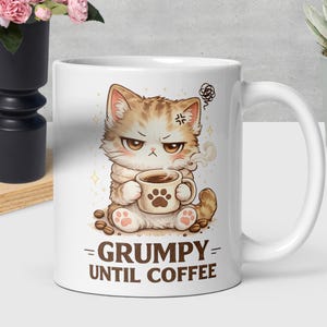 Op de afbeelding: Witte keramische mok met een chagrijnige cartoonkat die een koffiekopje vasthoudt. De kat is bruin en wit met een boze uitdrukking. De mok heeft de tekst "= GRUMPY = UNTIL COFFEE" erop gedrukt. Koffiebonen liggen verspreid.