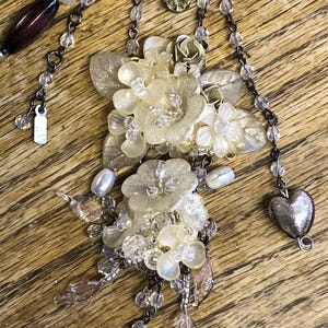 May include: A delicate necklace featuring a cluster of cream-colored, translucent flowers and leaves. The necklace includes clear beads, a silver heart charm, and a teardrop-shaped green gem. The chain is dark gray, and the necklace is set against a wooden surface.