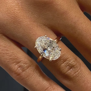 May include: A rose gold ring featuring a large, oval-cut diamond. The diamond is clear and sparkling, set with four prongs. The ring band is a simple, polished rose gold.