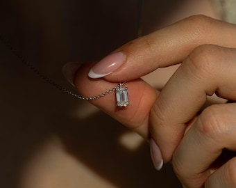 May include: A silver-toned necklace featuring a single emerald-cut clear gemstone pendant. The rectangular stone is set in a simple metal frame and hangs from a delicate, thin silver-colored chain, reflecting light with a clear, faceted appearance.