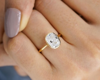Könnte beinhalten: Ein Goldring mit einem einzelnen klaren Edelstein im Kissenschliff, der in einer glatten Zargenfassung sitzt. Die Ringschiene ist dünn und poliert und unterstreicht das Design des facettierten Steins. Der Ring wird an einem Finger mit hellem Nagellack getragen.