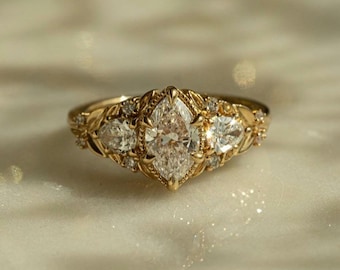 May include: A gold ring featuring a central marquise-cut diamond flanked by two pear-cut diamonds. The band is crafted with intricate metalwork detailing, including small round accent diamonds set within a leaf-like pattern along the shank.