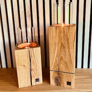 Könnte beinhalten: Zwei Kerzenhalter aus Holz mit Glaszylindern. Die Kerzenhalter sind aus hellbraunem Holz mit sichtbarer Maserung und natürlicher Oberfläche gefertigt. Jeder Halter hat einen quadratischen Sockel und einen zylindrischen Glaseinsatz zur Aufnahme einer brennenden Kerze.