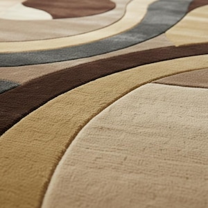 A high-angle shot of a rectangular hand-tufted wool area rug featuring a modern abstract pattern in brown, beige, tan, and grey. The rug is placed on a light oak wood floor in a bright living room next to a cream sofa, potted plants, and a wooden coffee table.