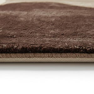 A high-angle shot of a rectangular hand-tufted wool area rug featuring a modern abstract pattern in brown, beige, tan, and grey. The rug is placed on a light oak wood floor in a bright living room next to a cream sofa, potted plants, and a wooden coffee table.