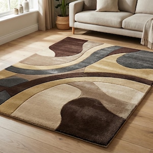 A high-angle shot of a rectangular hand-tufted wool area rug featuring a modern abstract pattern in brown, beige, tan, and grey. The rug is placed on a light oak wood floor in a bright living room next to a cream sofa, potted plants, and a wooden coffee table.