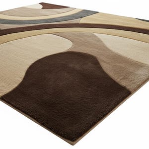 A high-angle shot of a rectangular hand-tufted wool area rug featuring a modern abstract pattern in brown, beige, tan, and grey. The rug is placed on a light oak wood floor in a bright living room next to a cream sofa, potted plants, and a wooden coffee table.