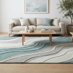 Op de afbeelding: Een woonkamer met een beige bank, houten salontafel en een rieten stoel. Een groot vloerkleed met een golfpatroon in blauw, grijs en wit domineert de vloer. De kamer heeft een maritiem thema, met een ingelijste foto aan de muur.