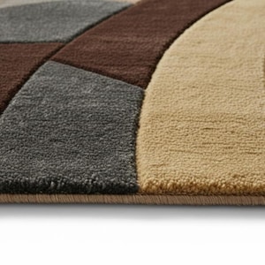 A high-angle shot of a rectangular hand-tufted wool area rug featuring a modern abstract pattern in brown, beige, tan, and grey. The rug is placed on a light oak wood floor in a bright living room next to a cream sofa, potted plants, and a wooden coffee table.
