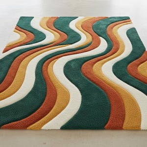 A high-quality Mid Century Modern hand tufted wool rug featuring a 3D abstract wave design in burnt orange, deep forest green, mustard yellow, and cream. The shag area rug has a plush multi-level pile and is styled on a wooden floor in a minimalist living room with vintage furniture.